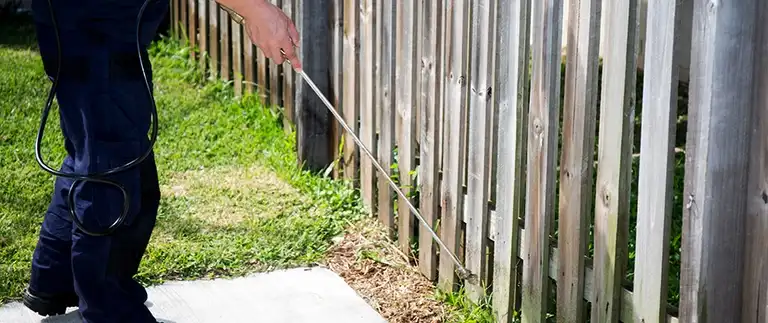 Technician spraying perimeter pest control treatment along a backyard wood fence line