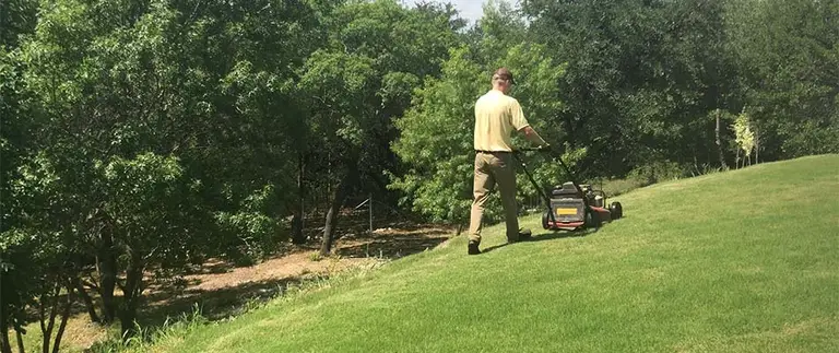 Lawn care technician mowing a grassy slope with a push mower near Fort Worth