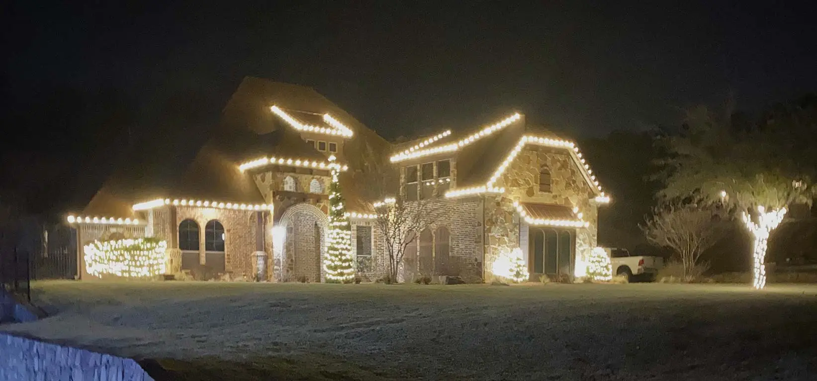 Warm white holiday lights outlining roofline, trees, and shrubs of Fort Worth home at night