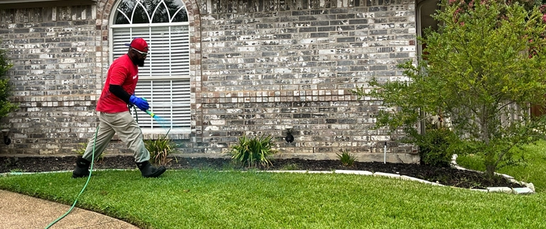 Technician spraying perimeter pest control along a brick home's foundation and landscaping in Lake Worth, TX