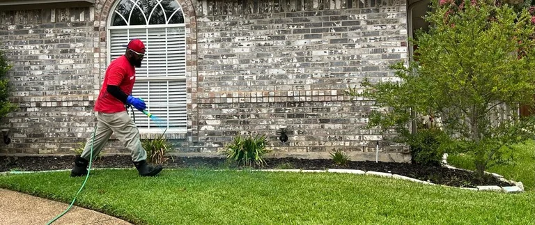 Technician spraying pest control treatment along the foundation of a Hudson Oaks brick home