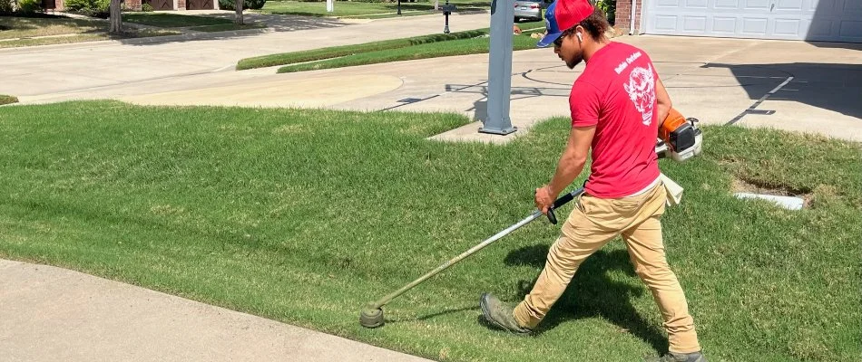 Worker in red shirt using a weed wacker on a lawn in River Oaks, TX.